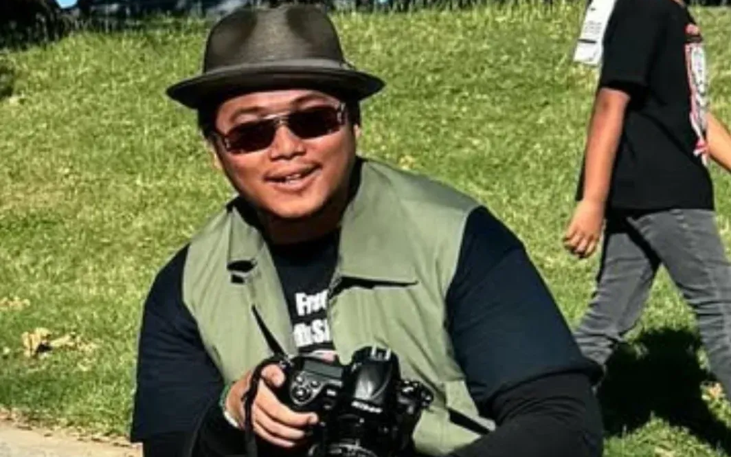 A smiling man in a porkpie hat, green vest, tan cargo pants, a thermal undershirt and a t-shirt holding a camera